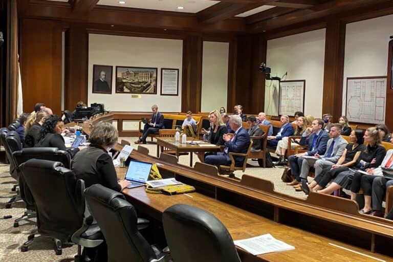 A hearing room in the Massachusetts State House with seats to the left for legislators and staff, a table in the middle for people testifying (in this case Kendalle Burlin O'Connell and Ed Coppinger), and then benches for guests to sit.